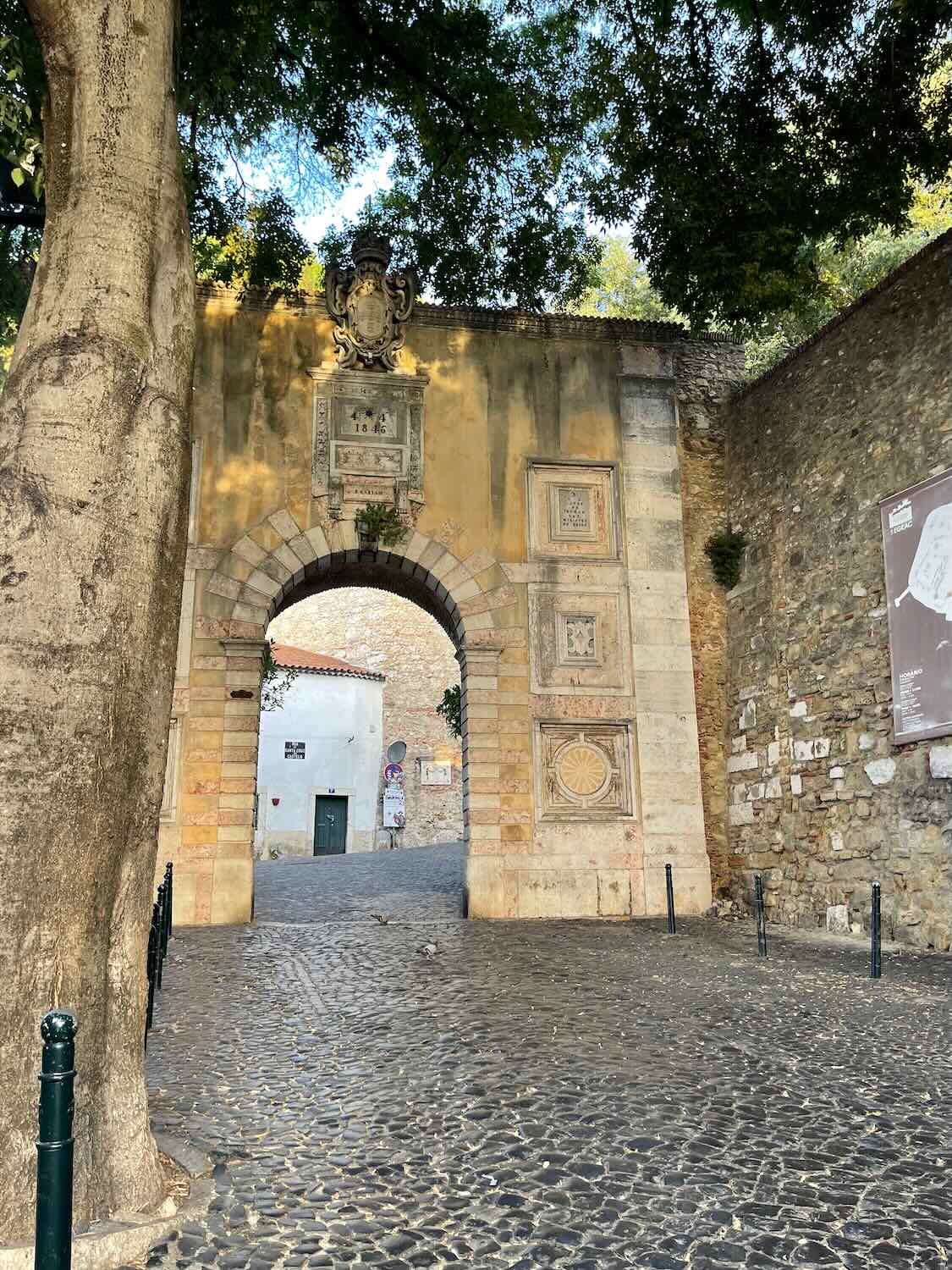 An ancient stone archway set into a tall wall with weathered carvings. A narrow, cobblestone street leads through the arch, and a large tree stands to the left, its branches reaching overhead. The street is quiet, with few people in sight.