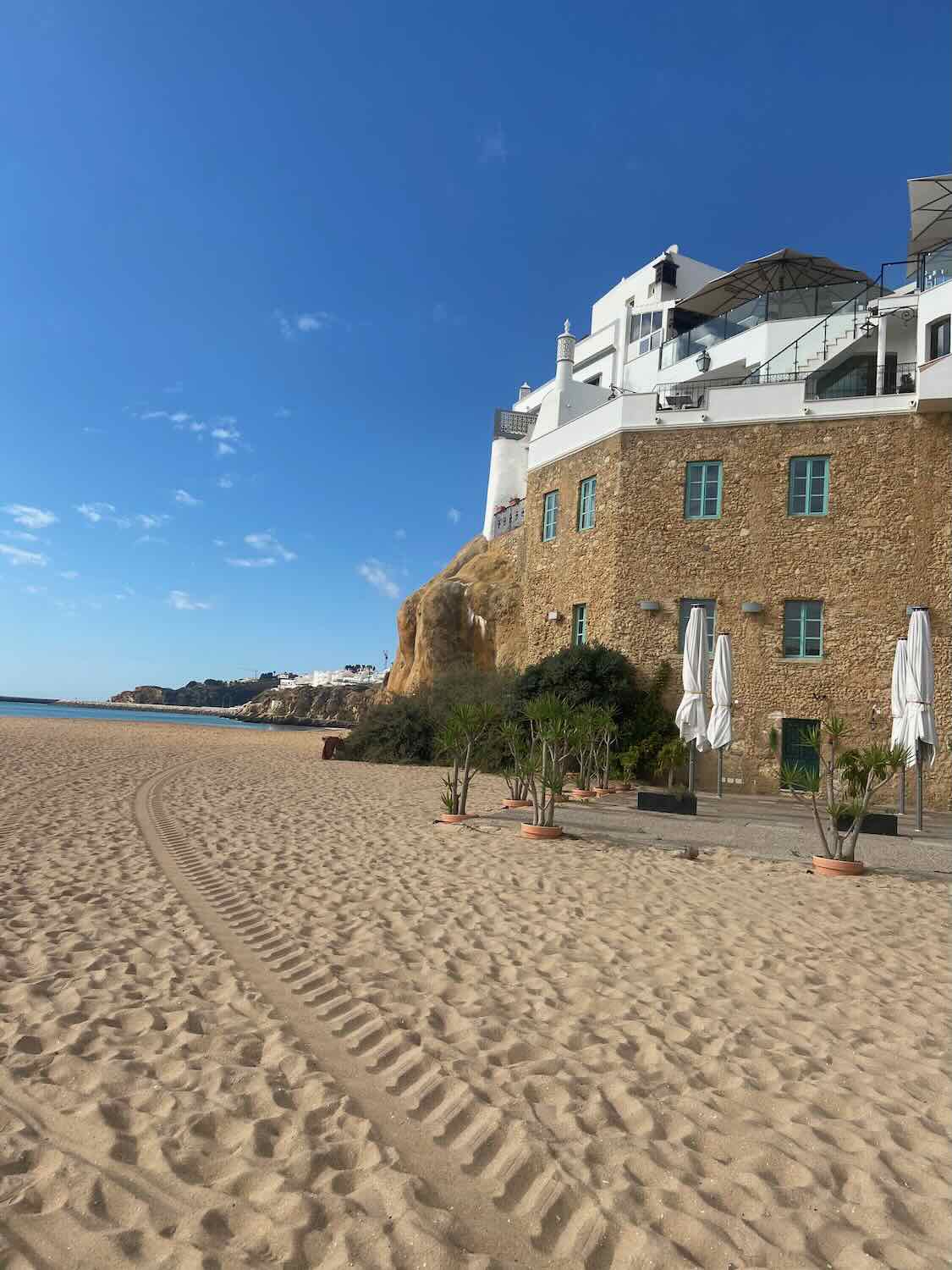 Pristine sandy beach in Faro, Portugal, with a clear blue sky, a unique rock formation, and white buildings atop, captured on a sunny day trip from Lisbon.