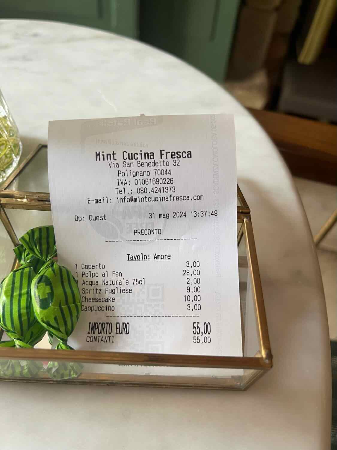 A close-up of a restaurant check from Mint Cucina Fresca in Polignano a Mare, showing a total amount of 55 euros for various items, including an octopus dish, water, Spritz Pugliese, cheesecake, and cappuccino. Two wrapped candies are also visible.