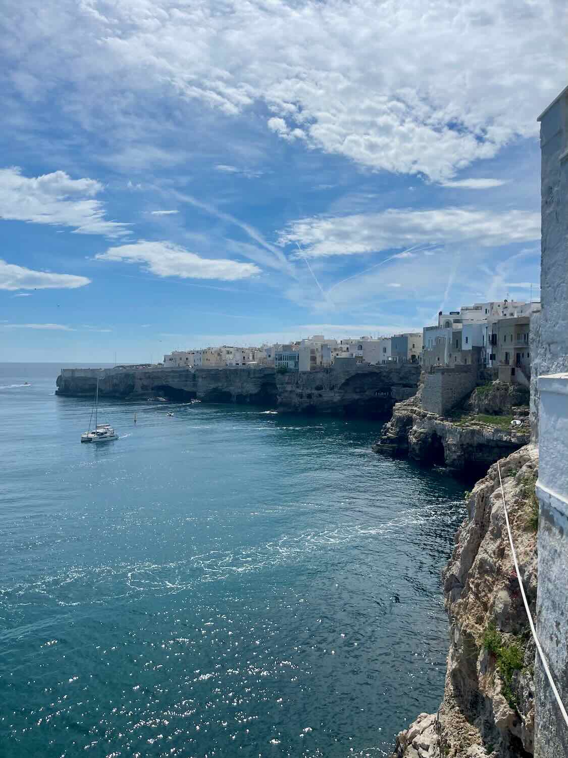 A stunning view of the coastline in Polignano a Mare, with clear blue waters, cliffs, and whitewashed buildings perched along the edge. A sailboat is anchored near the shore under a bright blue sky.
