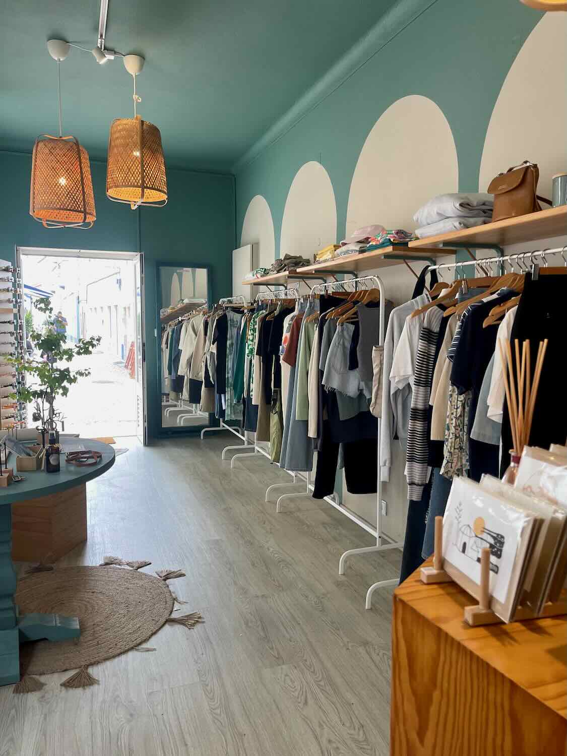 Interior of a clothing store with a teal ceiling and large wicker pendant lights. Clothes are neatly displayed on white racks along the teal and white arched wall. The floor is a light wood, and there is a circular woven rug near the entrance.
