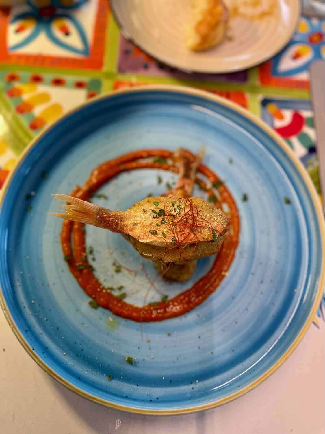 A beautifully plated fish dish with a decorative sauce swirl on a blue plate, representing a fine dining experience in Taormina.