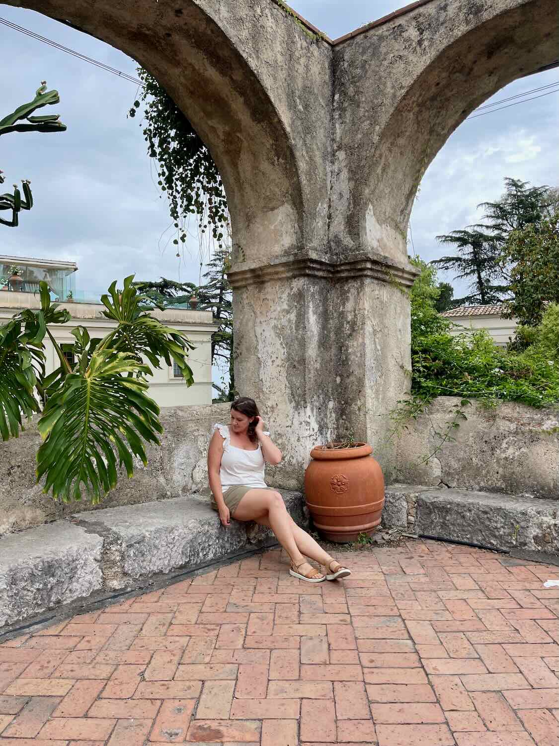 A woman in a white top and khaki shorts sits on a stone ledge under a weathered archway in Taormina, Italy. She is surrounded by lush greenery, and a large terracotta pot is beside her. The background features more vegetation and a cloudy sky.