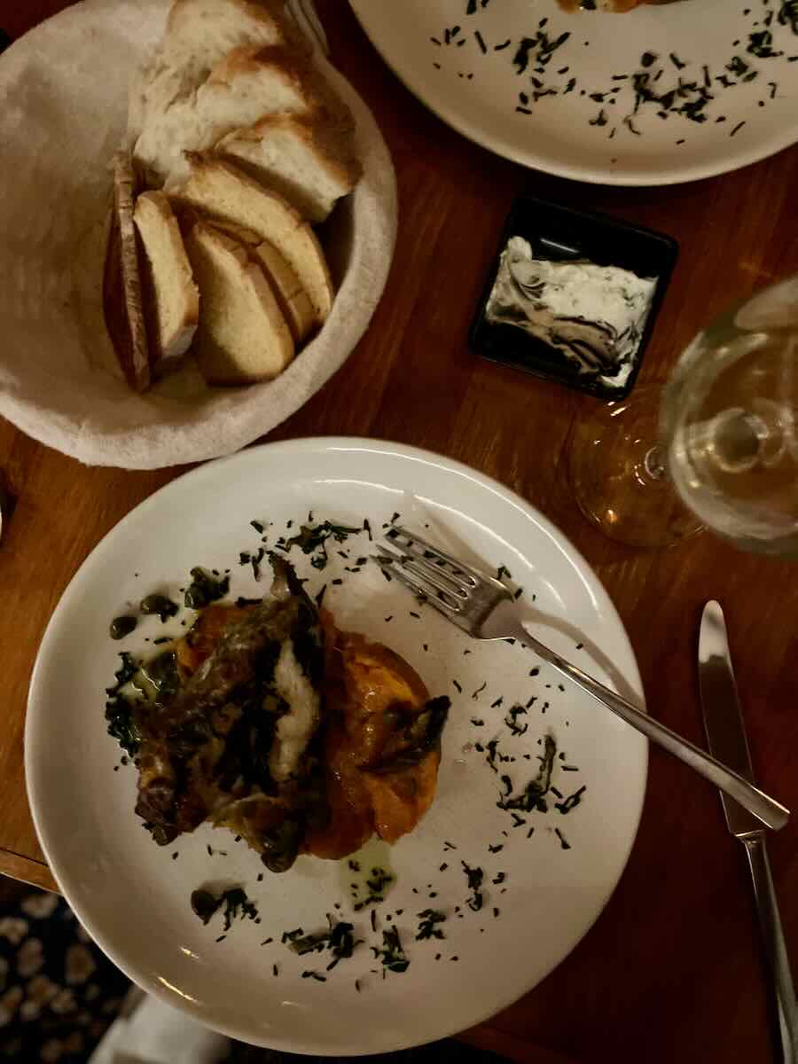 A dinner plate with a carefully plated dish of meat topped with vegetables, surrounded by a light drizzle of herbs. Next to the plate is a basket of sliced bread, a small dish of spreadable butter, and a glass of wine on a wooden table, set for a cozy meal.