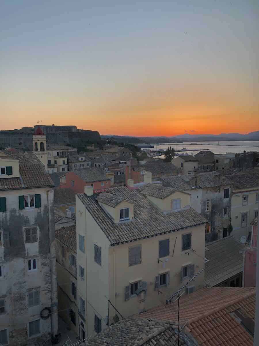 A scenic view of Corfu at sunset, featuring a blend of traditional stone-tiled rooftops, a church with a red bell tower, and the silhouette of a historic fortress overlooking the calm waters and distant hills under a golden-orange sky.