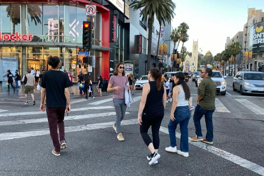 People crossing a busy city street with shops, palm trees, and a church in the background.