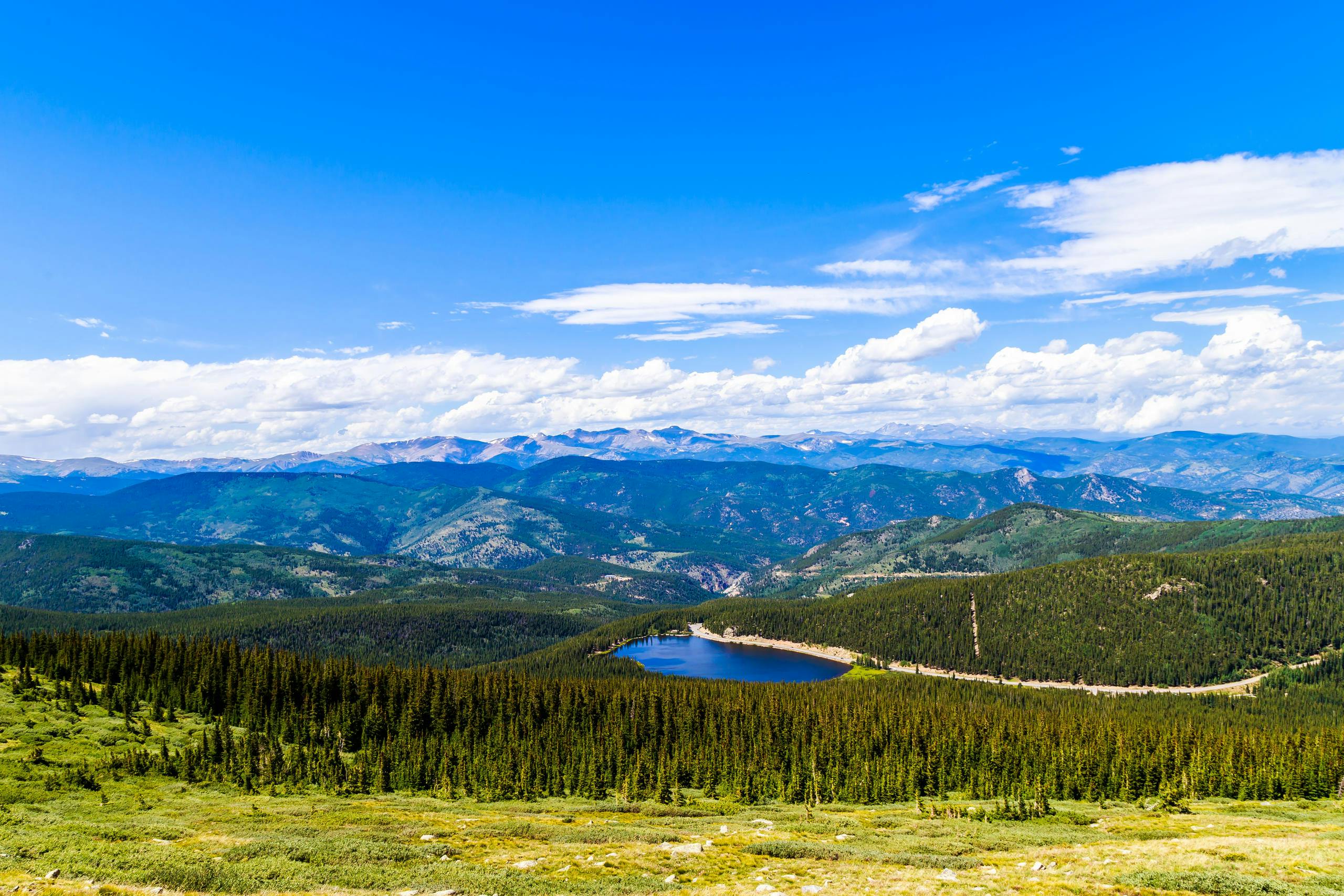 A scenic view of a forested valley with a small lake, surrounded by distant mountains under a clear blue sky.