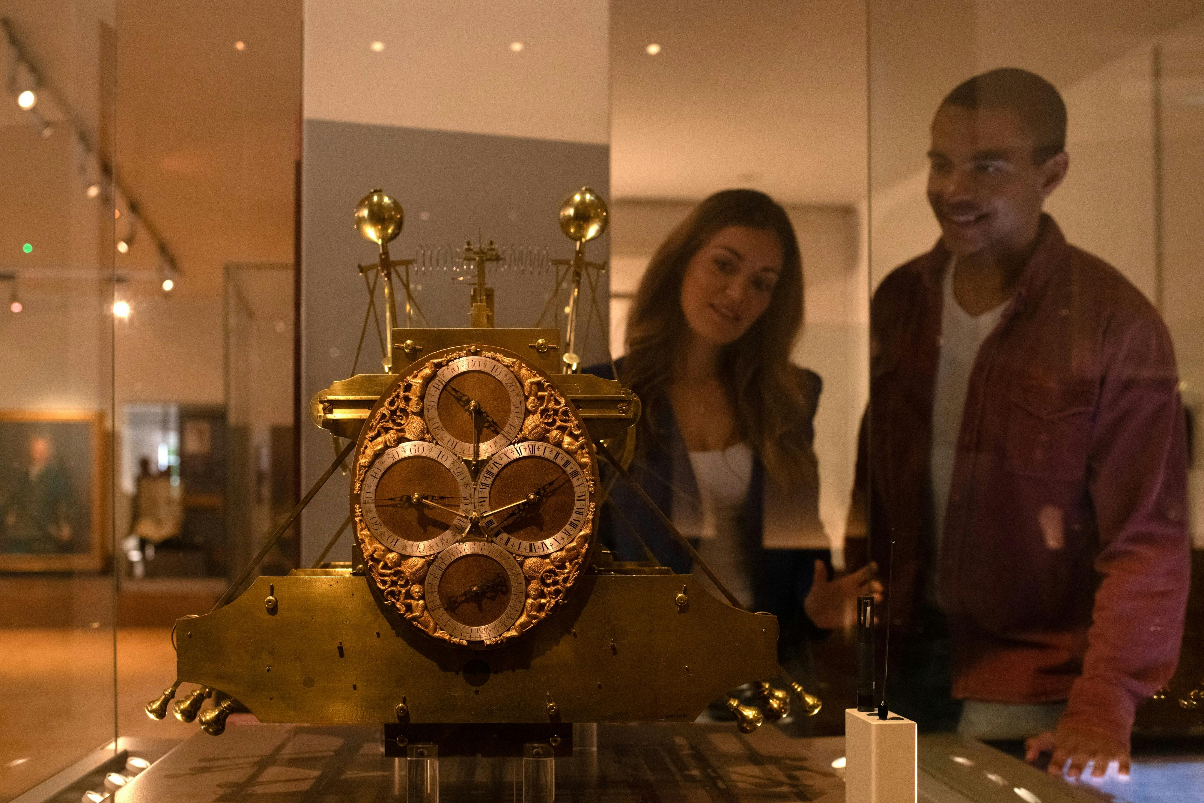 Two people smile while looking at an intricate antique clock in a museum display case.