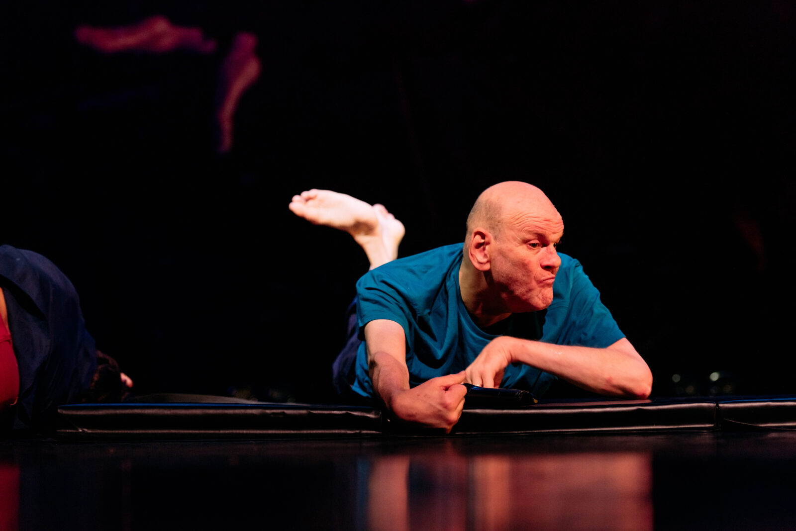 A bald man lies on his belly on the stage. He has one foot raised behind him.