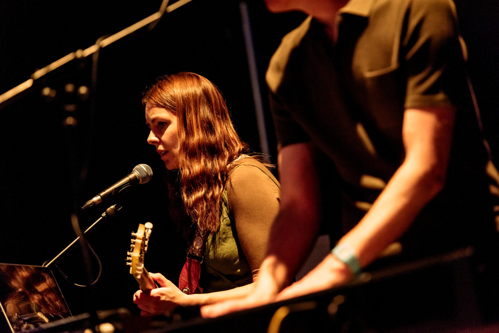 A close up shot of a woman playing guitar and singing. She appears to be in her late twenties, she has long wavy brown hair. We can see the torso of a man beside her.