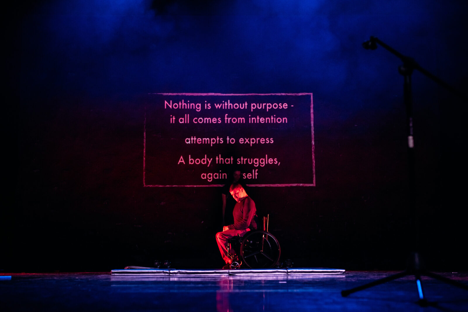 A man sits in his wheelchair at the back of the stage, there is brief glimpse of a woman behind him. Above his head there is a projection that reads 'Nothing is without purpose- it all comes from intention, attempts to express. A body that struggles against itself.'
