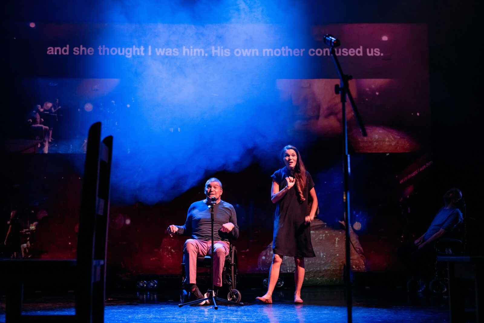 A man and a woman on stage. The man is sat down with a microphone in front of him and appears to be talking. He appears to be middle aged and is wearing a long sleeved blue top and pink trousers. The woman next to him is standing and she appears to be signing. Above their heads, there is a projected caption that reads 'and she thought I was him. His own mother confused us.'