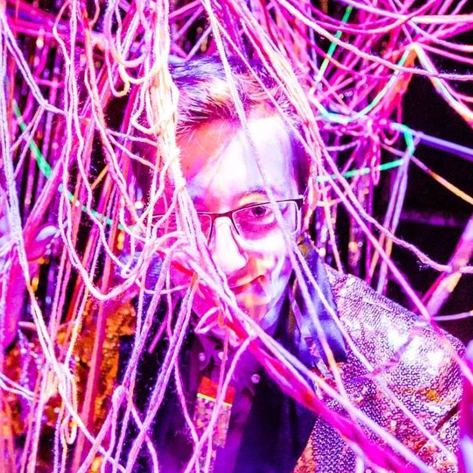 A photo of Sean Gallagher. The picture is washed purple and his face is slightly obstructed by various thanging threads. We can see that he has short hair and is wearing glasses.