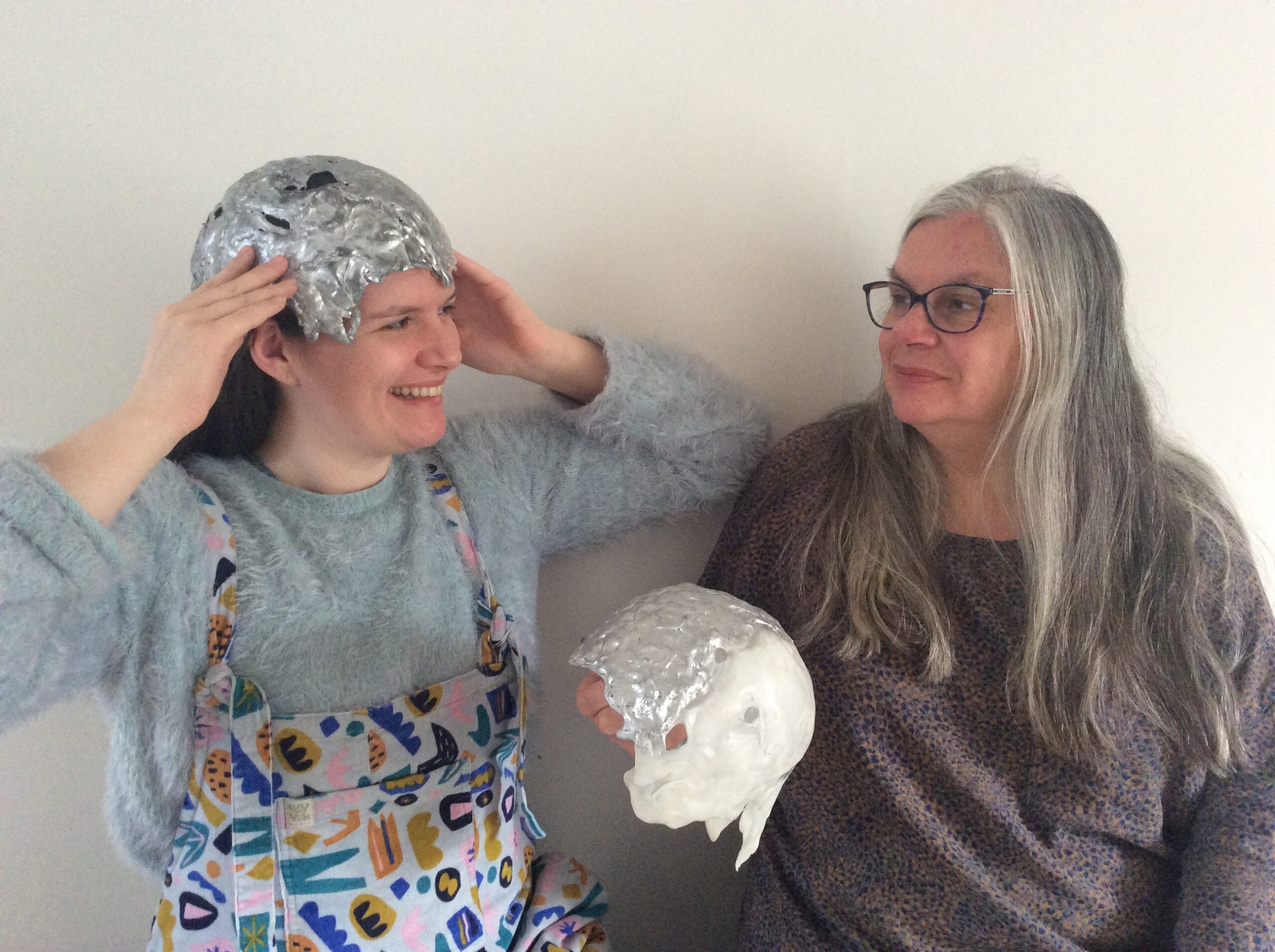 A young woman and an older woman sit together, looking at each other. In their hands they hold slivery sculptures that look like casts of their own heads.
