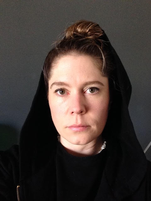 Bryony is a woman in her 30s with her hair tied back and wearing a black hoodie with the hood up. She stares intently at the camera.