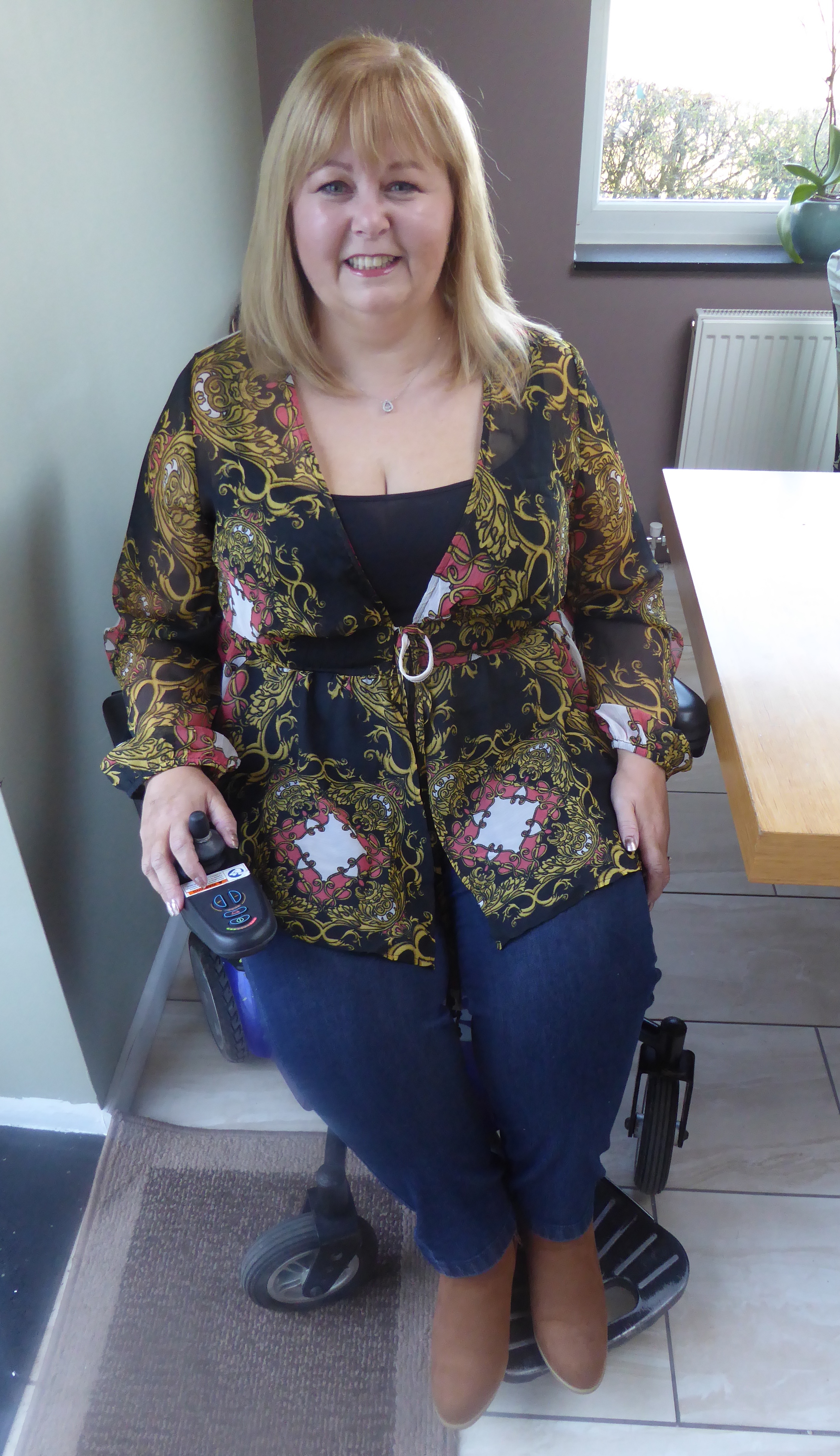 Lynley poses in her wheelchair wearing a scarf print kimono, tan boots and jeans