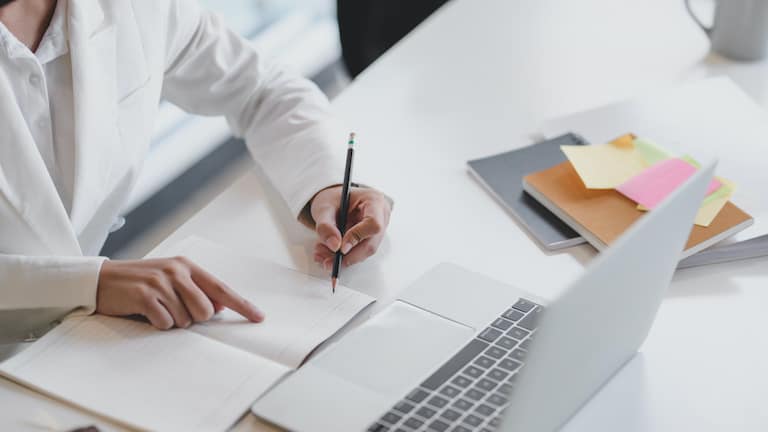 Cropped shot of businesswoman writing on notebook while working on laptop with office supplies on white table