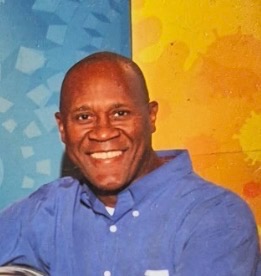 Obituary portrait of Bobby Lane. The casual photograph shows a Black man with a shaved head smiling broadly at the camera. He is wearing a blue button-down shirt over a white undershirt, positioned against a bright blue and yellow abstract patterned background.
