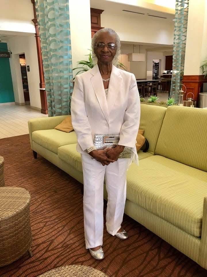 Portrait of an elderly woman wearing an elegant white suit jacket and pants with silver accessories, standing inside a lobby area.