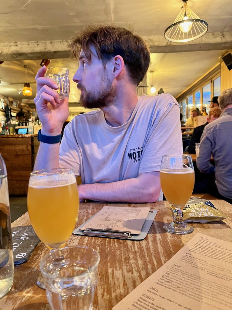 A man sits at a table holding a glass of water with two pints of hazy craft beer in front of him