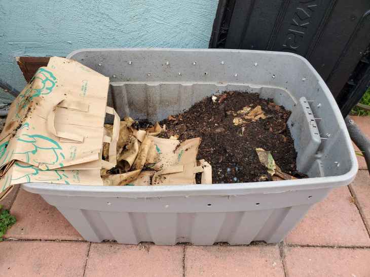 Eine graue Tasche, die für einen Wurmbehälter verwendet wird. Löcher sind gebohrt in die tote um die top 3 zoll der tote für luftstrom. Die Tasche enthält eine Mischung aus Material, aus dem die Wurmbettung besteht, Gussteile, und Essen. Einige braune Papiertüten sind an der Seite der Tasche, die verwendet werden, um die Oberseite des Bettenmaterials zu bedecken, wenn sie nicht verwendet werden.