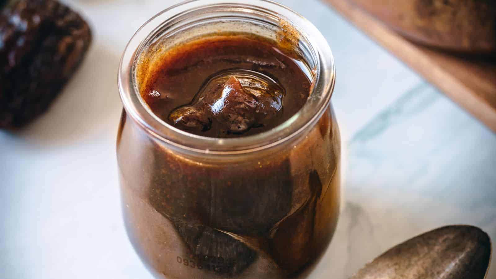 A glass jar filled with dark brown liquid, possibly homemade date syrup with a melting ice cube, resting on a light surface with a blurred background.