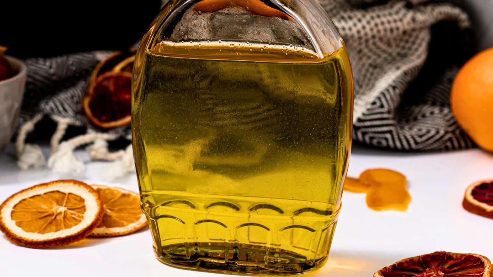 A glass jar of golden liquid, possibly oil, with textured sides. in the background, slices of dried citrus and a draped gray fabric enhance the vibrant, rustic setting.