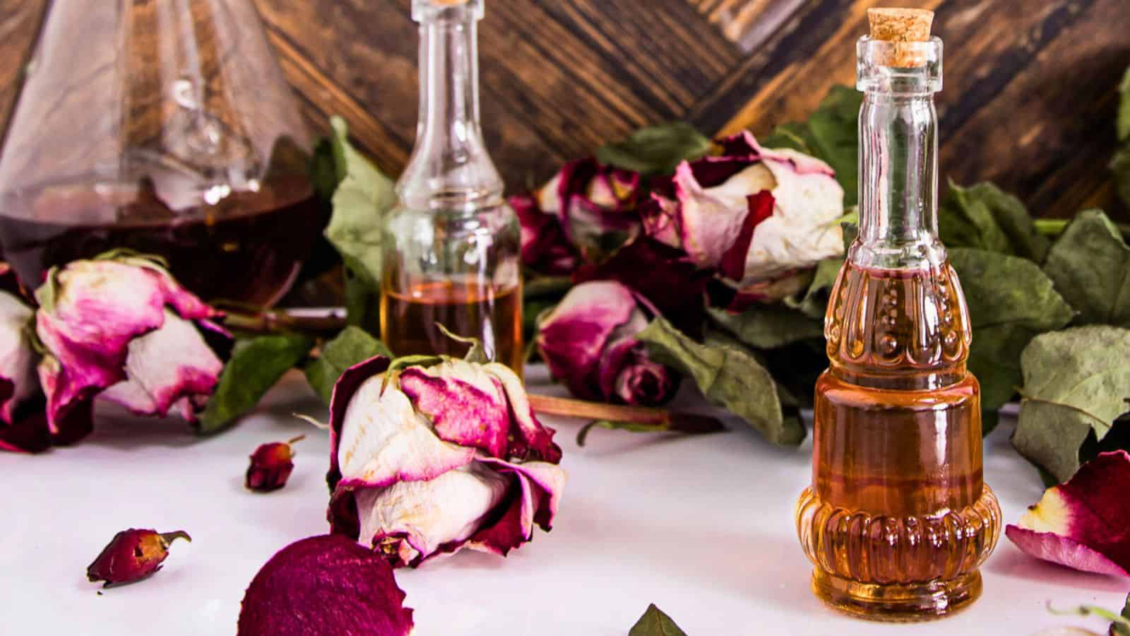 An assortment of glass bottles containing liquids on a wooden surface, accompanied by dried rose flowers and petals scattered around.