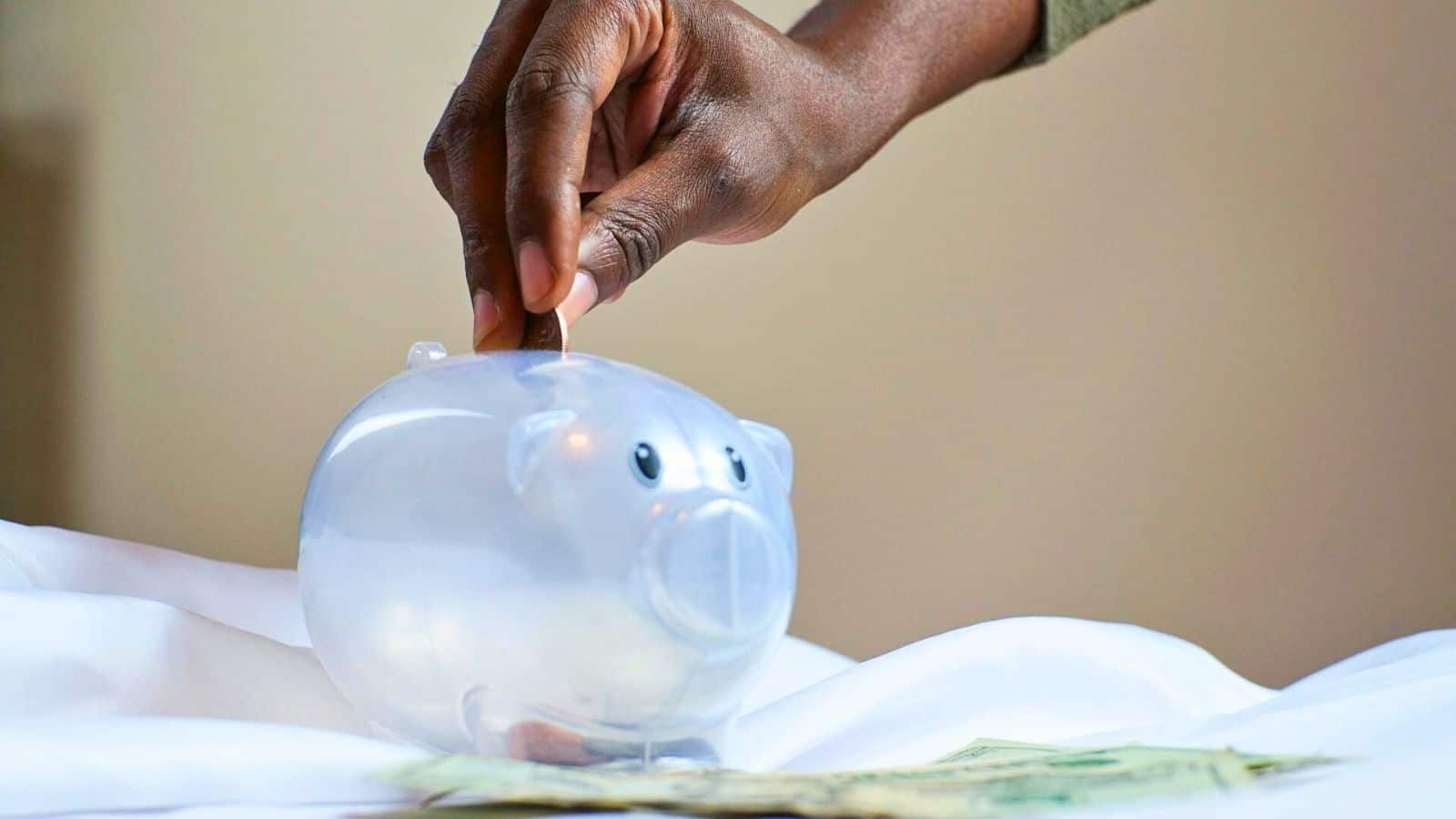 A person is placing a coin into a transparent piggy bank. The piggy bank is positioned on a white surface, and there are some dollar bills scattered around it. The background is neutral and blurred.