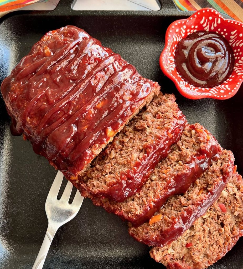 Overhead view of smoked meatloaf with silver fork and sauce in red cup on the side
