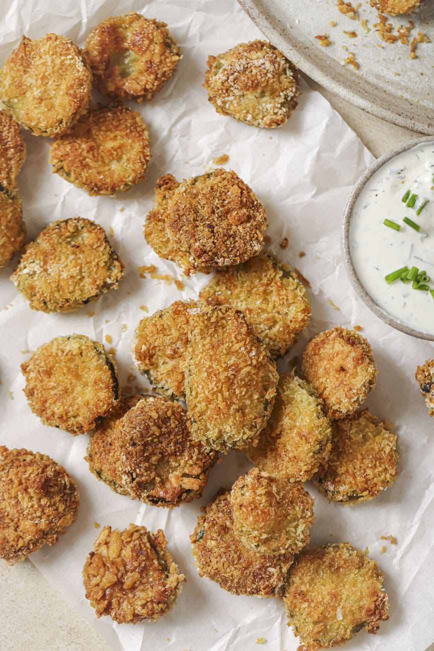 Golden breaded pickle chips on parchment with a bowl of ranch.