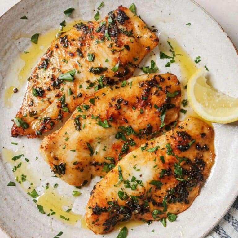 Three air fried tilapia filets topped with parsley on a white plate next to lemon wedges.