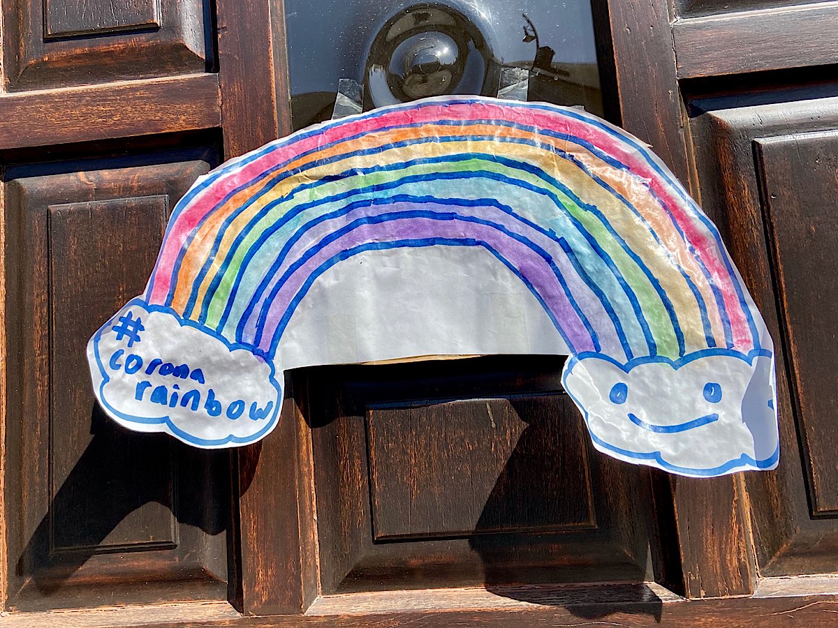 A rainbow that's cut out of paper and pinned onto a door. One end of the rainbow is in a cloud that says '#coronarainbow ' and the other side has a smiley face in a cloud