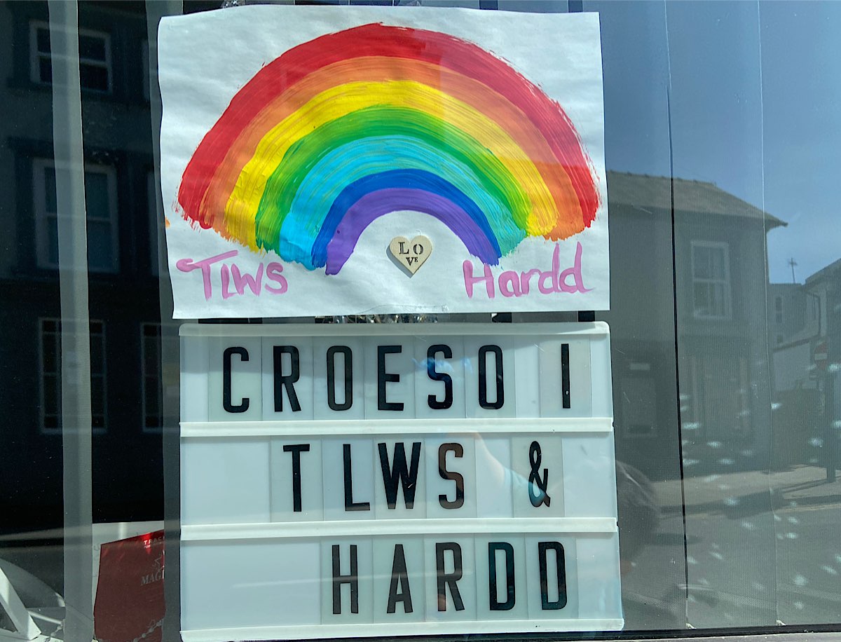 A rainbow in a window that's reflecting the town in the background. The rainbow has writing below that says 'Tiws Hardd' and a small wooden heart in the middle that says 'love'. Below the rainbow a leterboard spells out Croesi I Tlws & Harrdd'