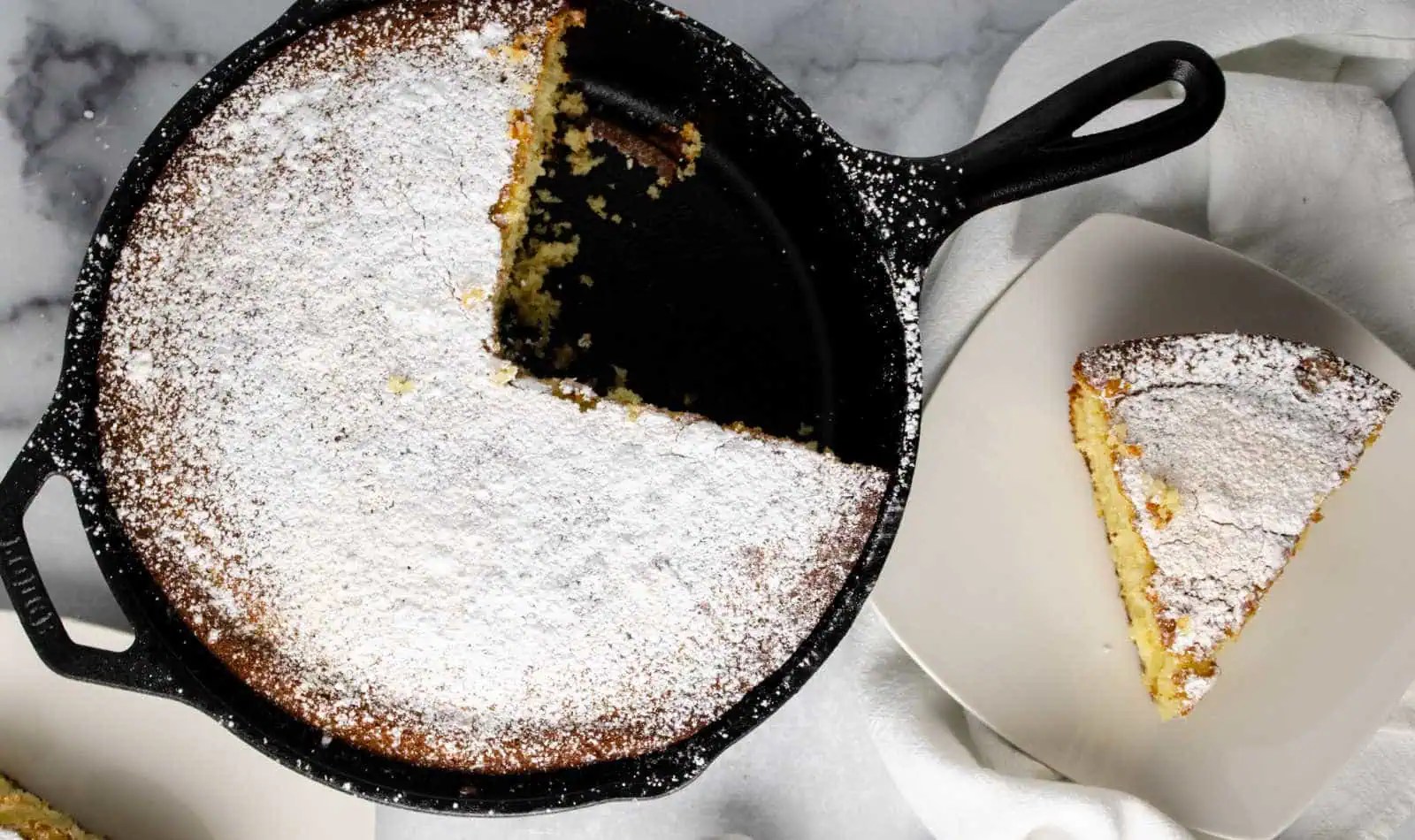 A cast iron skillet with a round, powdered sugar-dusted cake with a slice missing; the slice is placed on a nearby white plate.