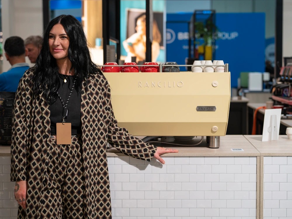 healthy brew hub nicole battefeld montgomery stands next to a rancilio espresso machine at a trade show