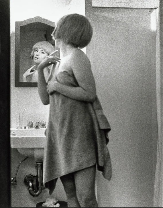 A person with short hair, wrapped in a towel, stands in a bathroom facing a sink. Their reflection is visible in the mirror, showing a thoughtful expression. The image is in black and white.