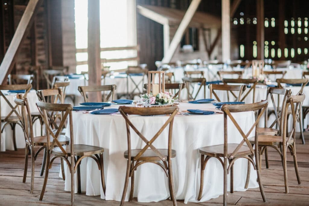 Wooden chairs surround round reception tables covered in white tablecloths in a rustic barn venue