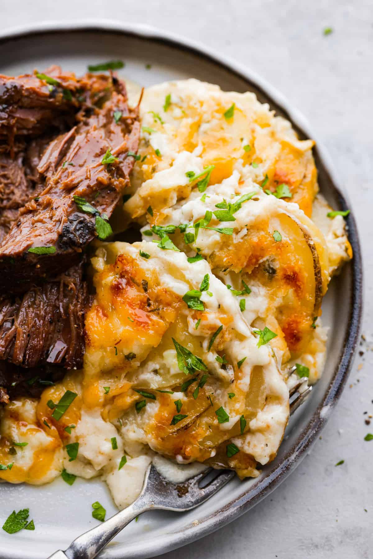 Creamy and cheesy au gratin potatoes are the perfect side dish to any comforting meal. Tender sliced potatoes baked in a rich and flavorful sauce with a golden layer of melty cheese on top. No one will be able to resist these heavenly potatoes. Close top view of au gratin potatoes on a gray plate served with shredded beef on the side.