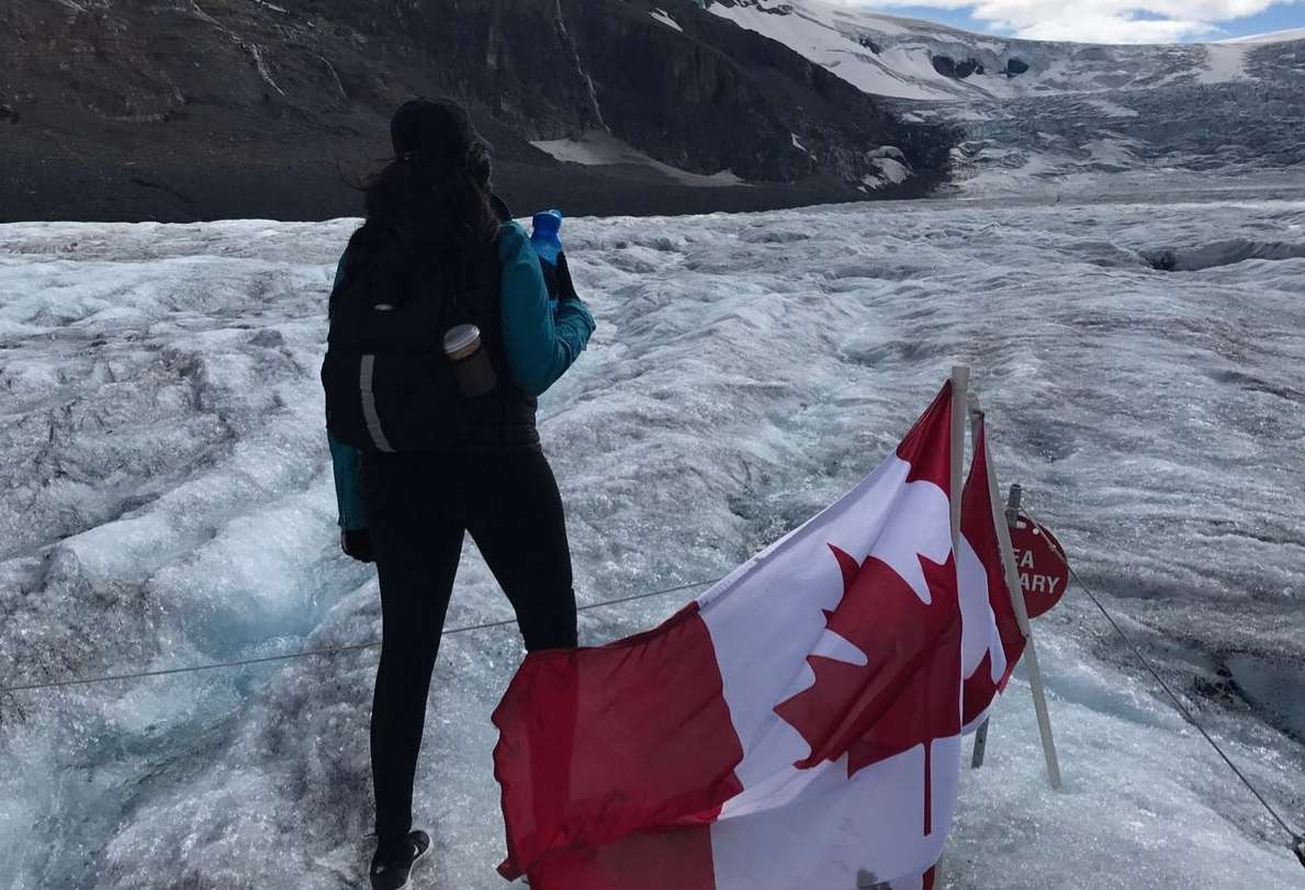 Woman wearing a backpack and jacket holding a water bottle, standing on a glacier by a Canadian flag looking at a mountain.