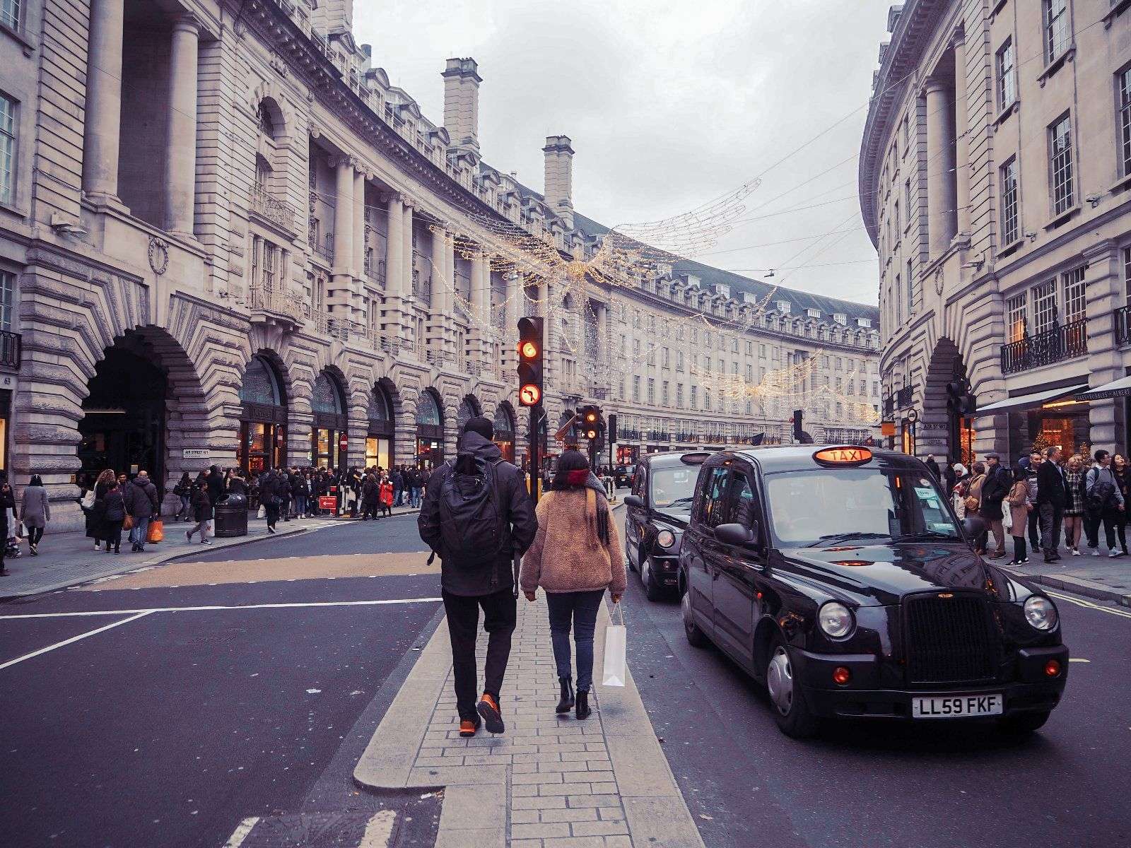 a woman and a man facing backwards and walking in the middle of the street with black cars on on the right side of them.