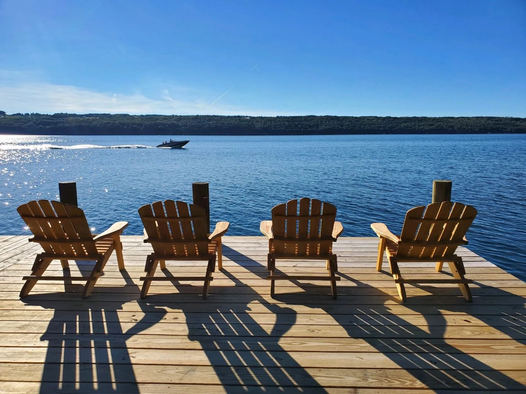 Wooden Adirondack chairs lined along a lakeside dock overlooking calm blue water with a boat passing in the distance.