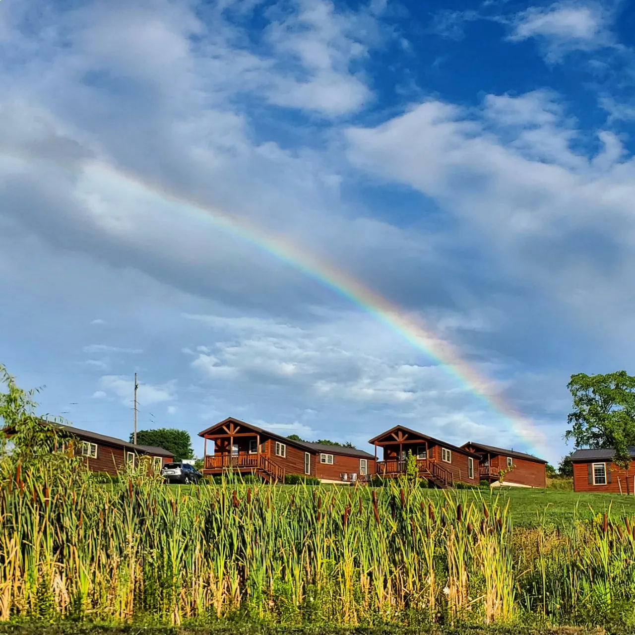 Rainbow stretching across the sky above a row of rustic cabins overlooking grassy fields and wetlands in the Finger Lakes region.