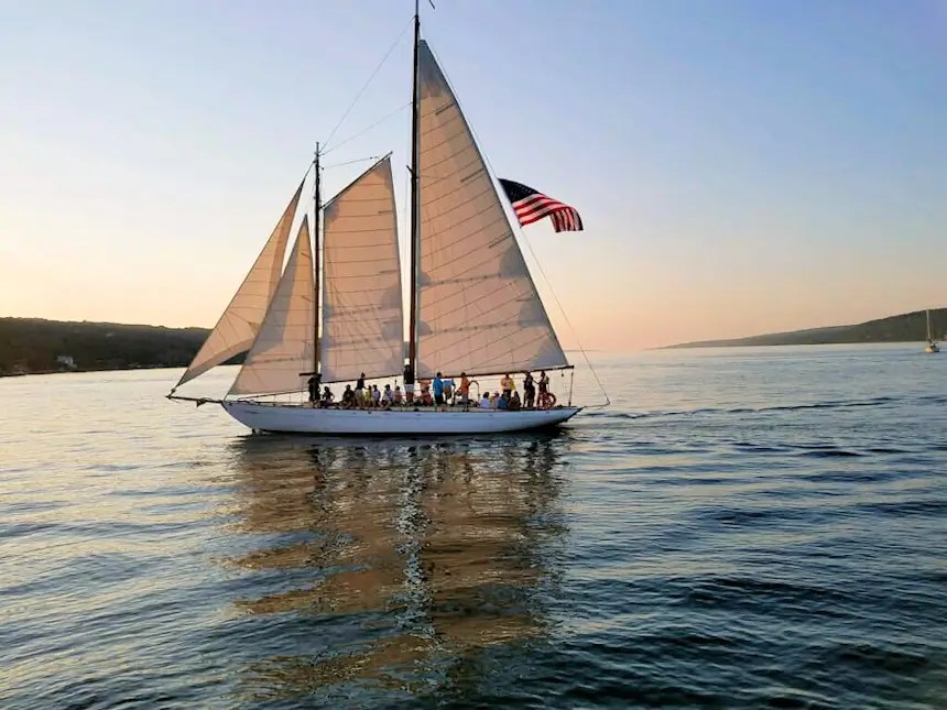 The Schooner True Love sailing across Seneca Lake at sunset with passengers aboard and an American flag flying from the mast.
