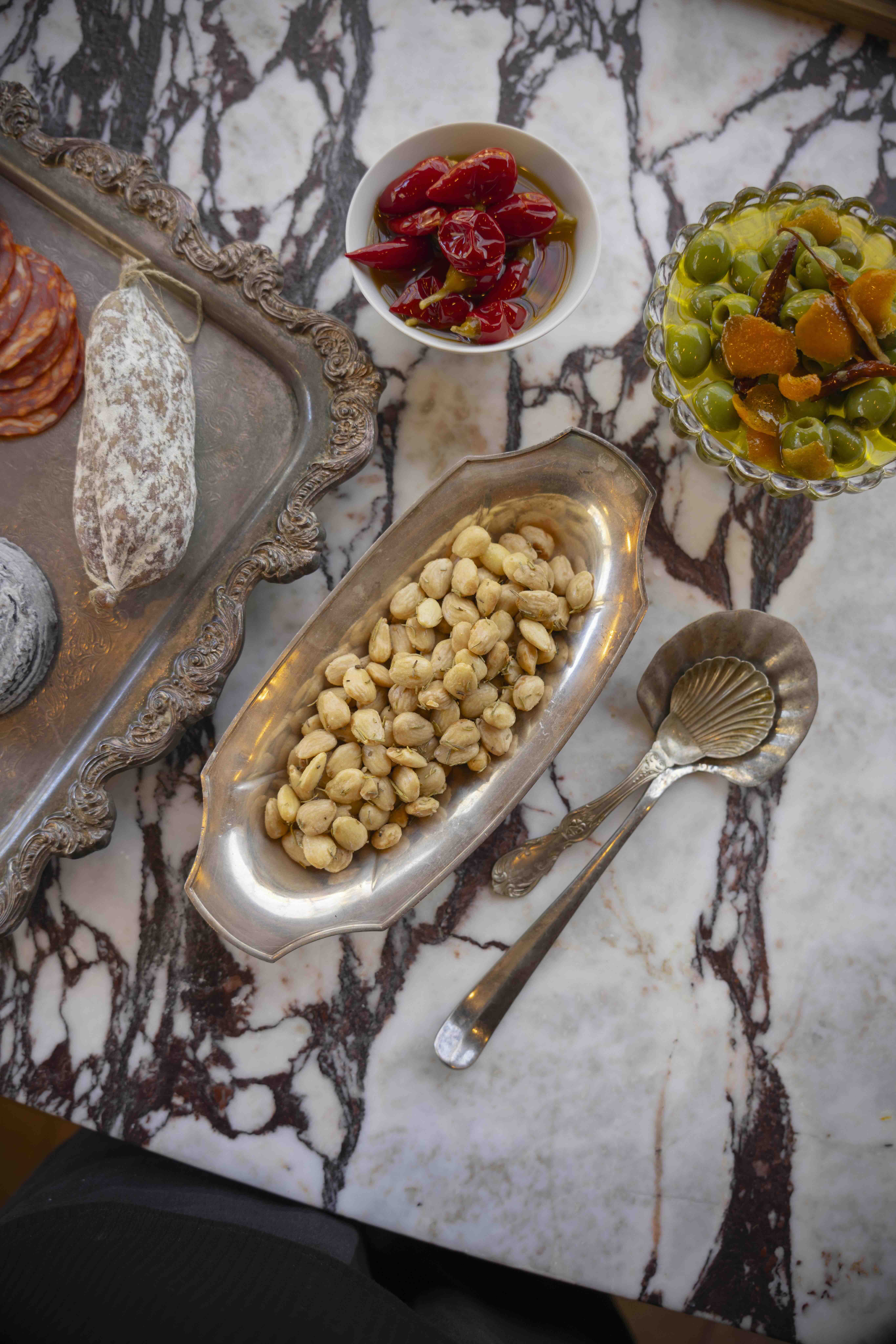 A variety of snacks are displayed on a marble table, including warm spiced nuts, salami, soft cheese, and olives.