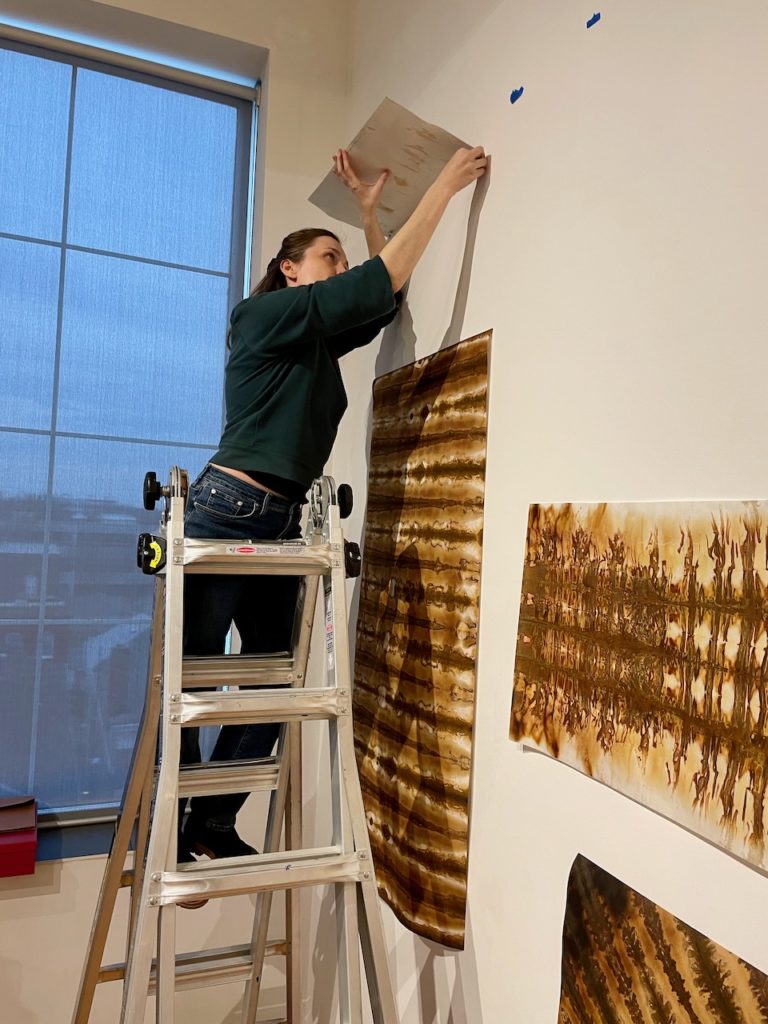 Betsy Zacsek pushing pins into a print in order to hang it on the wall. she's standing on a ladder