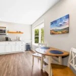 Bright kitchen with white cabinets, wooden floor, dining table set, and a landscape painting on the wall.