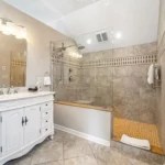 Modern bathroom with tan tiles, white vanity, and built-in bathtub.