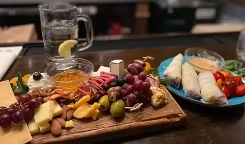 A charcuterie board featuring various cheeses, meats, nuts, olives, grapes, and a small bowl of honey, accompanied by a plate of spring rolls and a glass of water with a lemon slice.