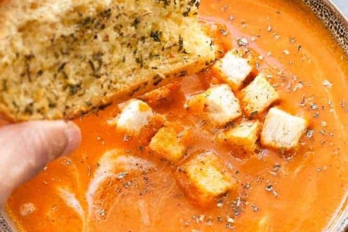 A hand holding a slice of bread in a bowl of tomato-based soup.